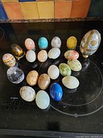 Overview of 21 decorative eggs made from various semi-precious materials on a dark glass surface