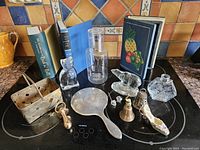 Full arrangement of all 15 decorative pieces including books, glass figurines, metal items, and glassware arranged on a black stovetop.