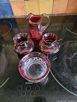 Four cranberry glass pieces arranged on a black stovetop: a pitcher, two vases, and a bowl.