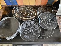 Photo of five clear glass serving platters on stove top, showing various shapes and designs including grape clusters and embossed figures.