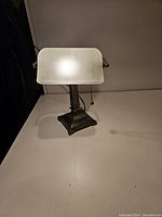 Front view of reproduction bankers lamp turned on, highlighting frosted hammered glass shade and metal base with pull chain.