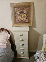 Bedside cabinet with six drawers and distinctive ceramic handles beneath a framed floral print hanging on the wall.