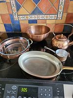 Full view of all five copper cookware pieces arranged on stove-top against colorful tile backsplash.