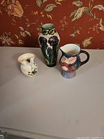 3 piece set of antique ceramics with floral designs displayed on a table against a floral wallpaper backdrop.