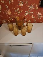 Full view of the lemonade service showing 1 jug and 4 glasses arranged on a white surface against floral wallpaper background.