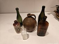Photo shows six antique bottles including green glass bottles, brown stoneware jug, and two glass medicine bottles with embossed text.