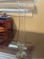 Full view of the tall slender glass bud vase on reflective surface, showing clear blown glass with etched decoration and scalloped top edge.