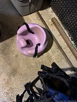 Pink infant booster chair placed on concrete floor with black safety straps attached.
