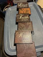 Photo of the vertical decorative wall hanging with four plaques featuring words Love, Faith, Hope, and plain plaque. Background is a blue plastic bin.