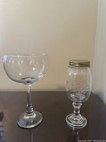 Side view of the two glasses showing the mason jar wine glass with a metal lid and the extra large wine glass on a table against a plain wall.