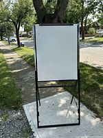 Front view of the portable whiteboard easel standing on concrete sidewalk with attached clip at top for paper pads.
