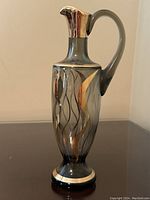 Front view of the vintage hand-painted glass oil decanter showing the decorative gold and silver wave patterns, curved handle and metallic spout on smoky gray glass.