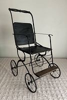 Side angle of antique collapsible stroller showing black fabric seat and backrest, metal frame, four metal spoked wheels, and wooden footrest.