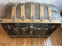 Front angled view of the antique metal and wood trunk showing embossed metal and wooden strips on the domed lid and front face.