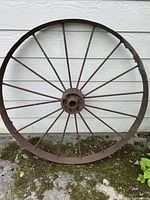 Full view of the antique iron cart wheel against a light wall and on mossy ground, showing overall condition and shape.