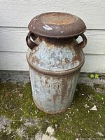 Full view of the large metal milk can with lid, showing rusted and weathered pale blue/gray paint and metal handles