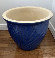 Blue ceramic plant pot standing on wooden floor, showing overall shape, blue exterior with leaf patterns and cream top rim.