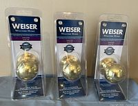 Three Weiser Welcome Home brass door knobs in original packaging displayed upright showing label and knobs.