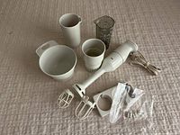 Braun hand blender main unit, two metal beaters, whisk attachment, opaque white and transparent measuring cups, additional small accessories in plastic bags spread on a beige textured surface.