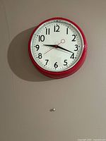Front view of the red framed round analog wall clock hanging on a wall, showing the face with numbers and hands.