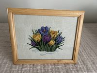 Framed cross stitch embroidery artwork depicting purple and yellow spring flowers in a wooden frame on a table.