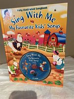 Colorful illustrated children's song book titled 'Sing With Me My Favourite Kids' Songs' with an embedded CD on cover showing farm animals and a barn.