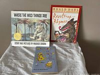 Three children's books shown on fabric surface: 'Where The Wild Things Are' hardcover, 'Revolting Rhymes' softcover, 'My Stepdad is an Alien' softcover.