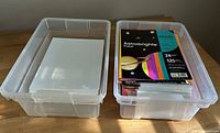 Two transparent plastic bins containing printer papers. One bin holds white cardstock or photo paper sheets and the other contains a pack of Astrobrights colored paper.
