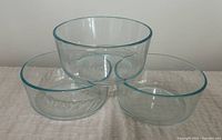 Three clear Pyrex glass bowls stacked and arranged on a white cloth surface, showing size and shape.
