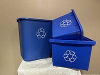 Photo showing three blue plastic recycling bins stacked and arranged, highlighting their shapes, colors, and recycle symbols.