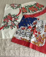 Three colorful vintage Christmas tea towels with festive holiday prints including bells, poinsettias, holly, winter houses, and floral patterns