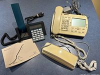 Three vintage telephones including Bang & Olufsen Beocom 1401 with manual, beige telephone with display and keypad, and beige telephone handset with power adapter.
