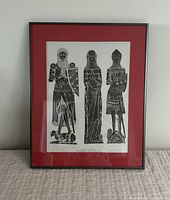 Framed vintage black and white brass rubbing print with red mat and black frame, showing three medieval figures.