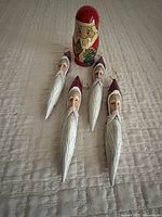Four vintage Santa head tree decorations and a Santa Matryoshka doll arranged on a beige quilted surface.