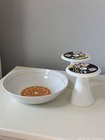 Photo of Mikasa Art Deco style white bone china candleholders with black, floral decorated tops next to a white ironstone bowl with orange floral pattern inside and gold rim.