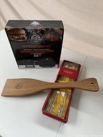 Photo of the three BBQ accessories: Pit Boss cover in original box, William Sonoma pineapple skewers in red box, and wooden BBQ cleaning spatula.