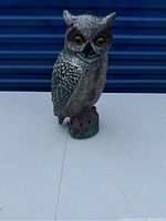 Photo of a plastic owl decoy on a white surface with a blue background. The owl is detailed with painted feathers and yellow eyes, sitting on a green-brown base.