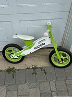 Photo of a green and white Rider kids balance bike with two green wheels, white seat and grips, shown standing outdoors against a garage door.