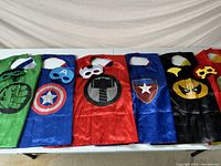 All six capes and six masks laid out on table.