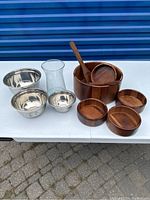 Image showing three wood salad bowls with three wooden spoons, three metal mixing bowls, and a clear glass vase arranged on a white table.