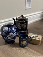 Overview photo showing five items: navy blue ceramic zodiac mug, navy blue floral vase, black floral teapot with wicker handle and base, and nested Japanese lacquer boxes.