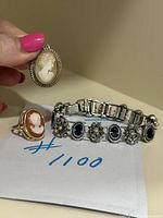 Photo of cameo pendant held in hand with detailed silver frame, a cameo ring with gold plated band, and a silver-tone costume bracelet with blue and rhinestone embellishments.