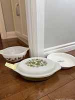 Three vintage dishes arranged on a floor: lidded casserole with green floral pattern on top, pie plate with blue Corningware logo, and a patterned mixing bowl.