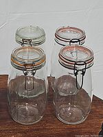 Four clear glass canister jars shown from above and side, highlighting glass lids with pink-orange rubber gaskets and metal wire clasps