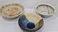 Three ceramic bowls shown together: swirled yellow & blue inside bowl, rust & cream geometric bowl, and blue & cream bowl with handles and spiral design.