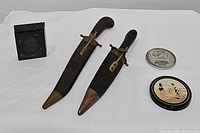 Full lot showing two daggers with scabbards, black etched box, metal lion coin, and small round Asian trinket.