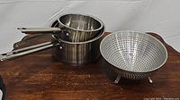 Photo showing two stacked Professional Gourmet stainless steel saucepans and vintage aluminum footed colander on wooden surface.