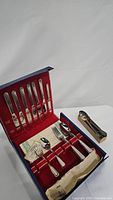 Flatware set open in blue storage case with red velvet lining showing six dinner knives, three pieces of flatware and assortment of 12 silver spoons in small wooden tray.