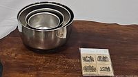 Photo of the four stainless steel nesting mixing bowls stacked inside each other on a wooden surface next to the sealed pack of vintage magnets.