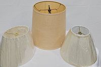 Three lamp shades arranged on white background, one large tan linen shade and two smaller white pleated shades covered in plastic